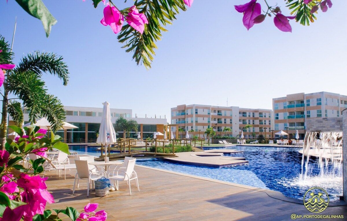 Área de piscina com espreguiçadeiras e guarda-sóis, um elemento aquático e um edifício com várias varandas ao fundo.