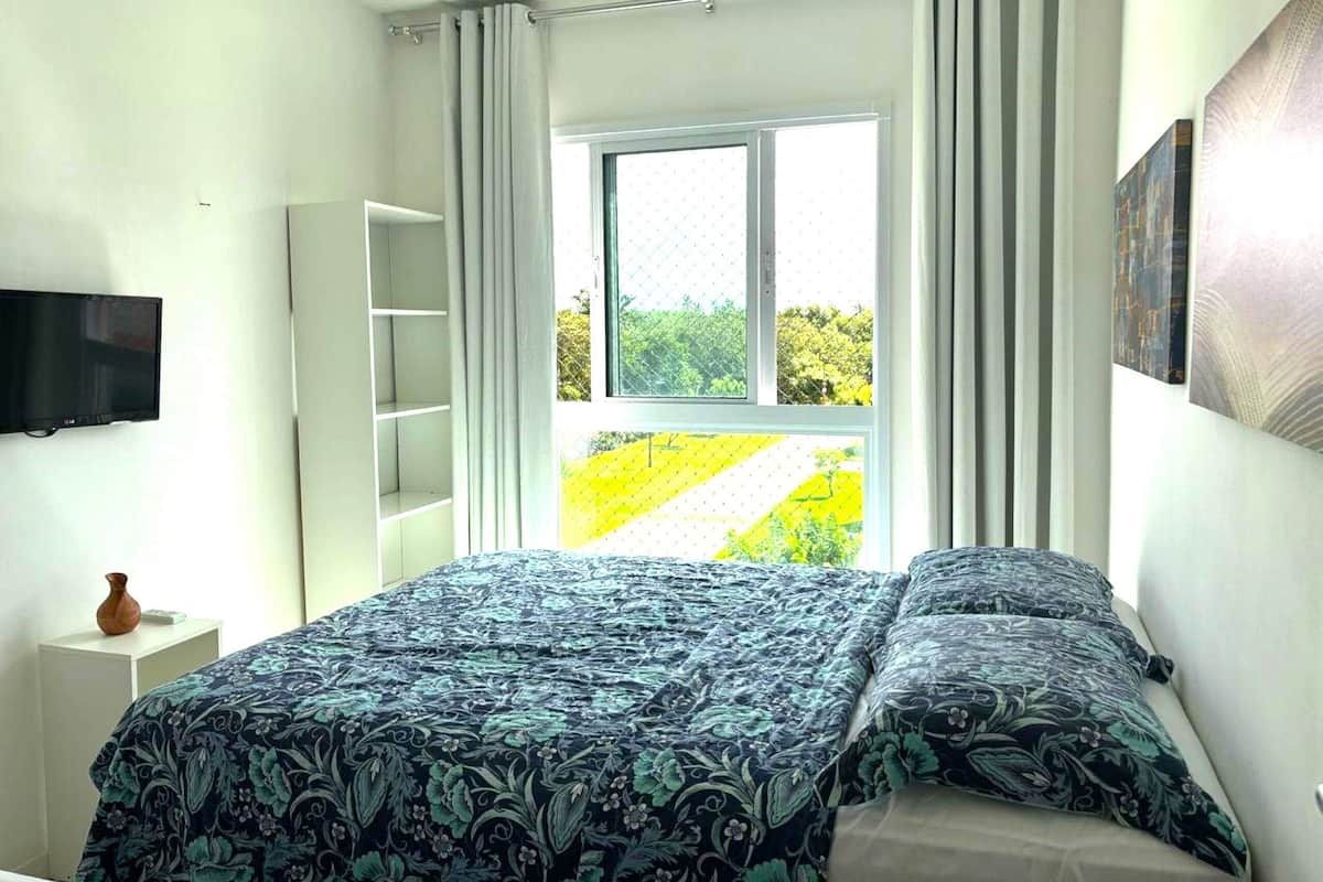 Une chambre à coucher avec un lit, une télévision, une fenêtre avec des rideaux et une vue sur la verdure.