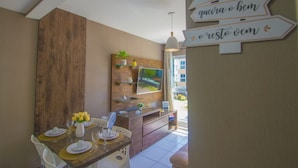 A dining area with a wooden table set for two, a TV mounted on the wall, and directional signs. - Flat em Porto de Galinhas - Muro Alto Clube (Ipojuca)