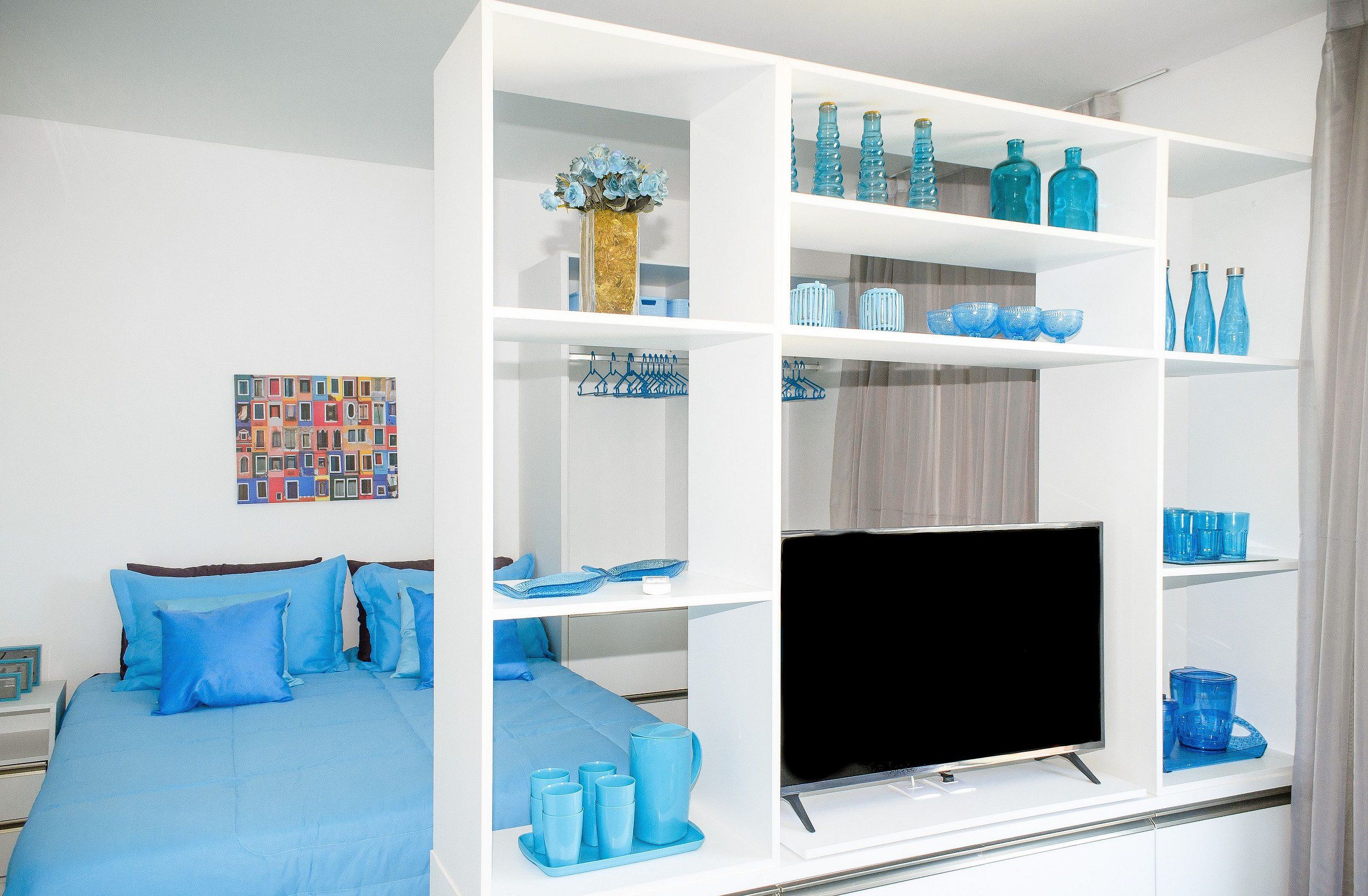 Een slaapkamer met een bed, een televisie en planken met blauwe spullen.
