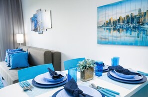 Un coin repas avec une table dressée pour deux personnes, des chaises bleues, un tableau représentant un paysage urbain et un canapé avec des coussins bleus.