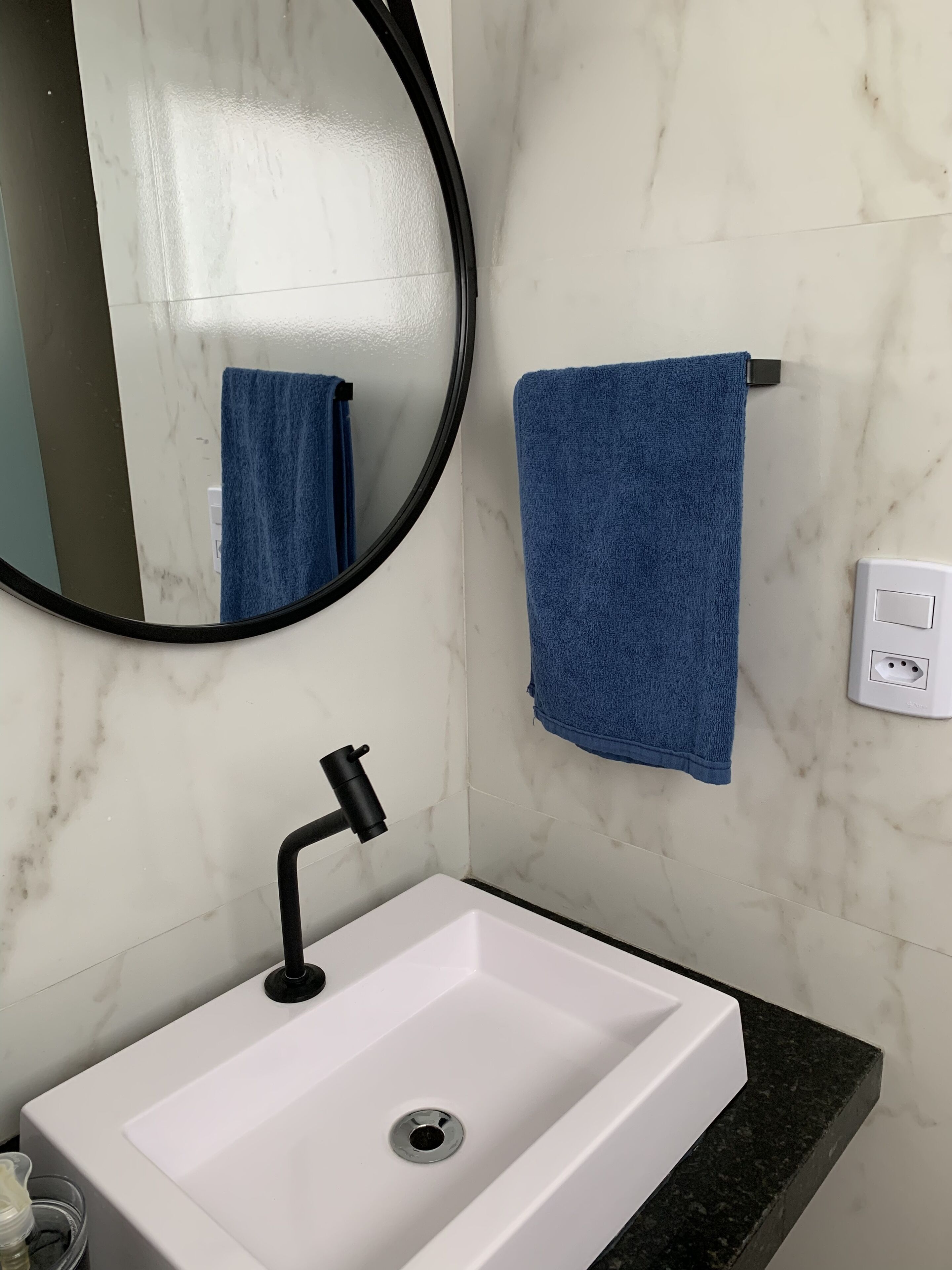 Un baño con un lavabo blanco, un grifo negro, un espejo redondo y una toalla azul colgada en un perchero.