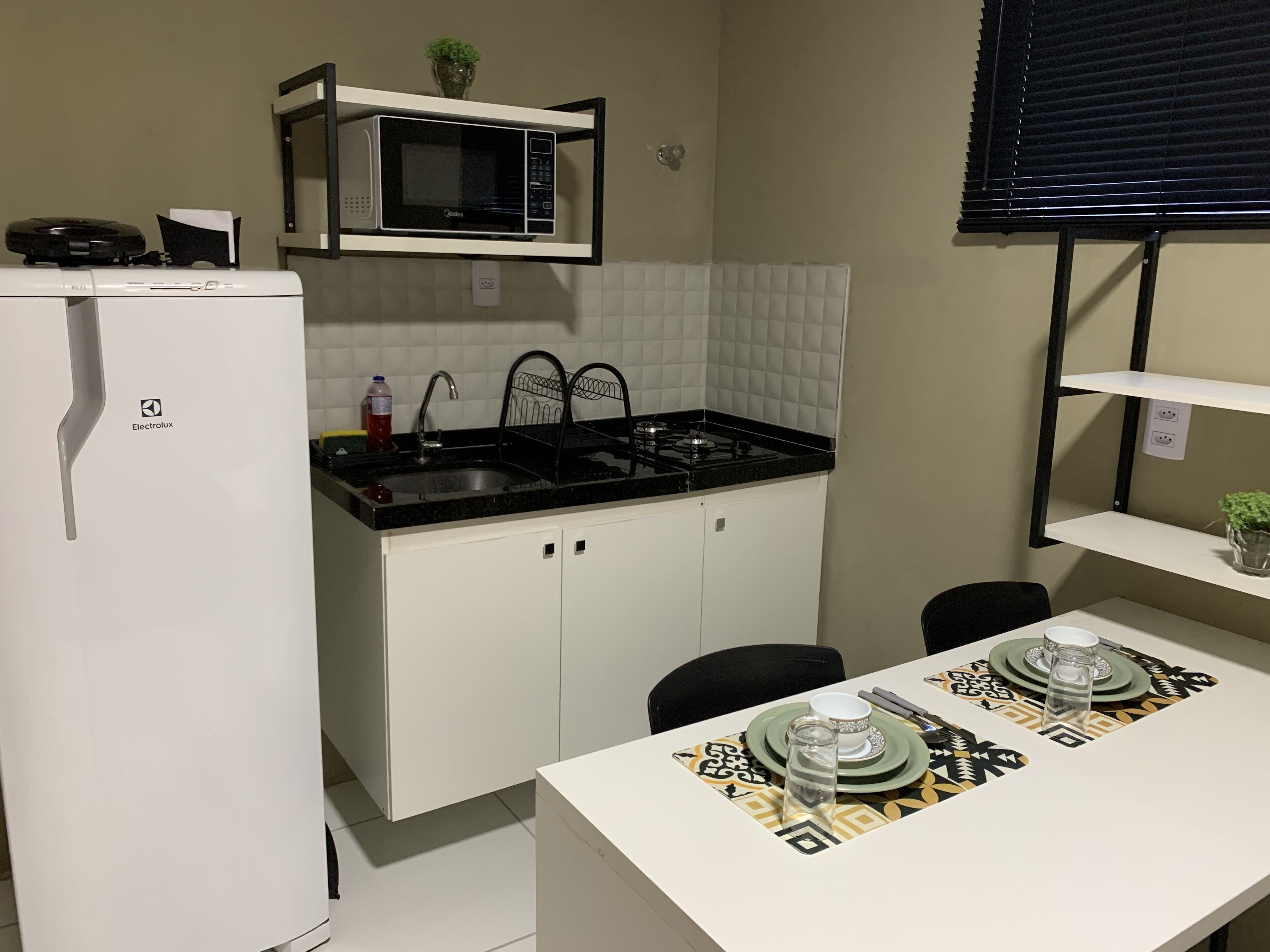 A compact kitchen with a white refrigerator, a microwave, a sink, and a stove. There is a dining table with a patterned tablecloth and a few dishes.