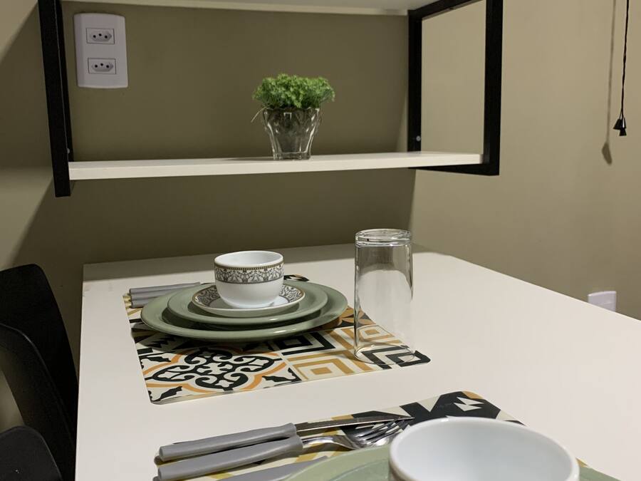 Una mesa de comedor con un juego de platos, una taza y cubiertos. Encima de la mesa, hay una pequeña planta en maceta sobre una estantería.