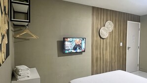 A hotel room with a bed, a television mounted on the wall, a towel stack, a wooden panel wall, and a door. - Edifício Creta (São Luís)