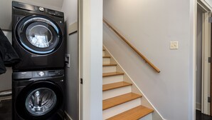 A laundry area with stacked washers and dryers, a wooden staircase, and a door leading to another room.