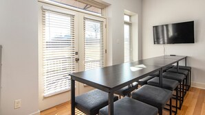A modern room with a long table and six chairs, a flat-screen TV mounted on the wall, and large windows with blinds.