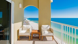 Ein Balkon mit zwei Korbstühlen und einem kleinen Tisch, mit Blick auf einen Strand mit klarem, blauem Wasser und einer fernen Küstenlinie.