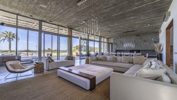 Amplia sala de estar con un sofá grande, una mesita de centro y un sillón. Cuenta con una chimenea de piedra y vistas al exterior a través de amplios ventanales.