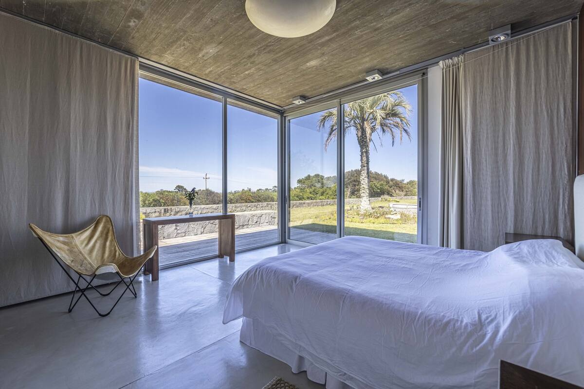 Quarto moderno com uma cama grande, uma cadeira e vista para palmeiras através de grandes janelas.