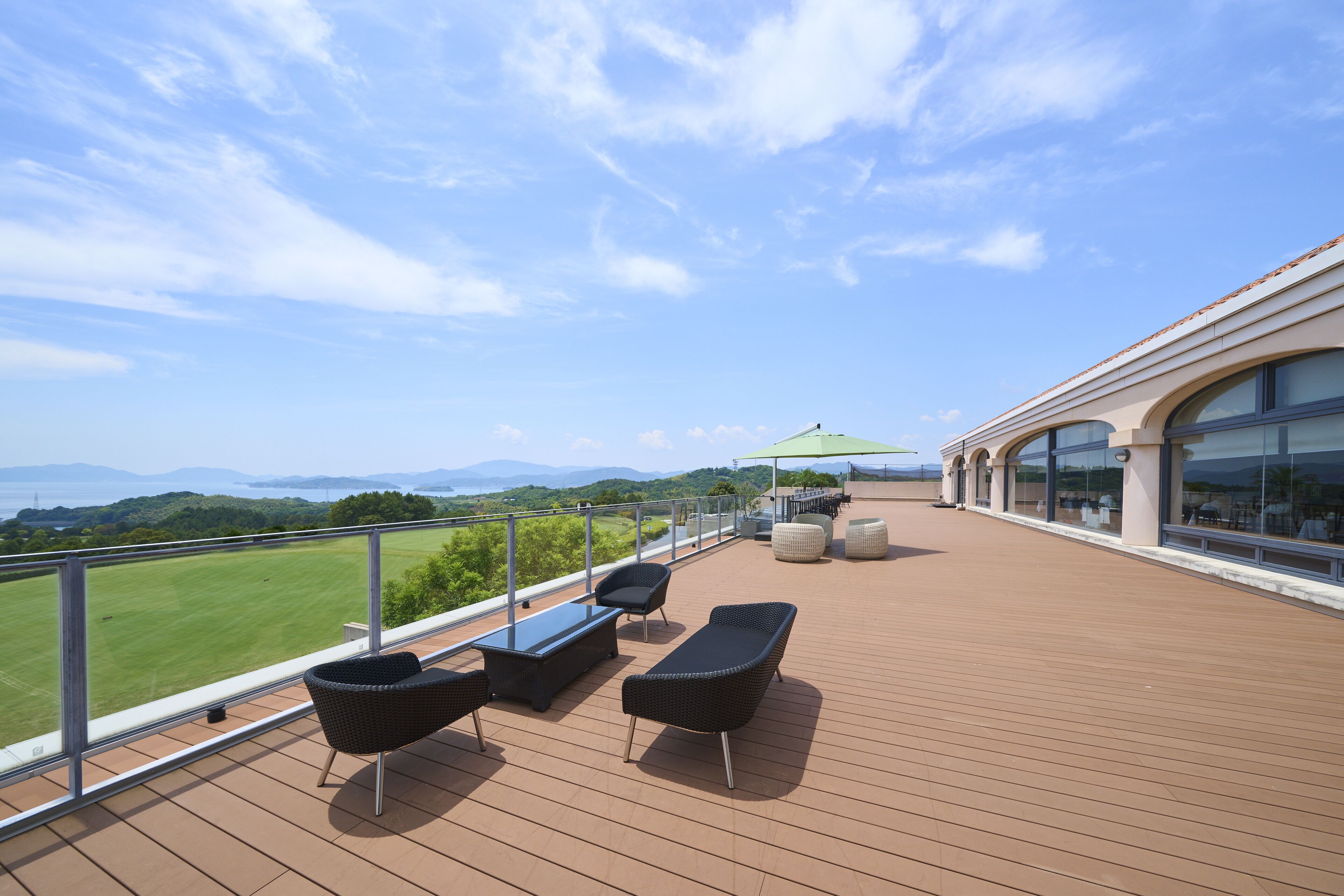 A deck with outdoor seating, a view of a green field, and a building with large windows.
