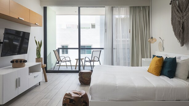 A modern hotel room with a large bed, a TV on a white cabinet, a balcony with chairs, and a view of buildings. - Opal Suites (Playa del Carmen)