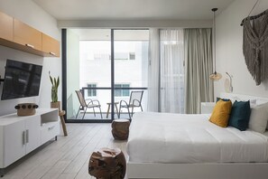Quarto de hotel moderno com uma cama grande, uma televisão sobre um móvel branco, uma varanda com cadeiras e vista para prédios.