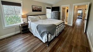 Ein Schlafzimmer mit Bett, Deckenventilator, Fernseher, Kommode und Fenster mit Blick auf Bäume.