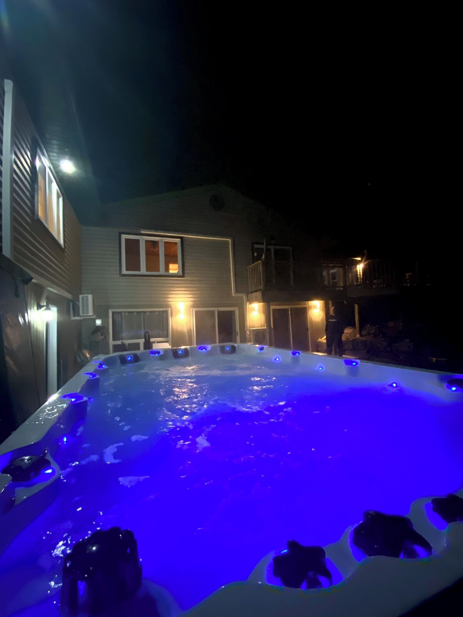 Un bain à remous doté d’un éclairage bleu, entouré d’une maison avec de nombreuses fenêtres et un balcon.