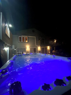 A hot tub with blue lighting, surrounded by a house with multiple windows and a balcony.