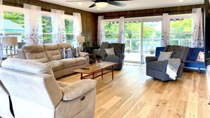 Un salon spacieux avec un canapé beige, un fauteuil gris et un canapé bleu. On y trouve un ventilateur de plafond, un plancher en bois et de grandes fenêtres avec des rideaux.