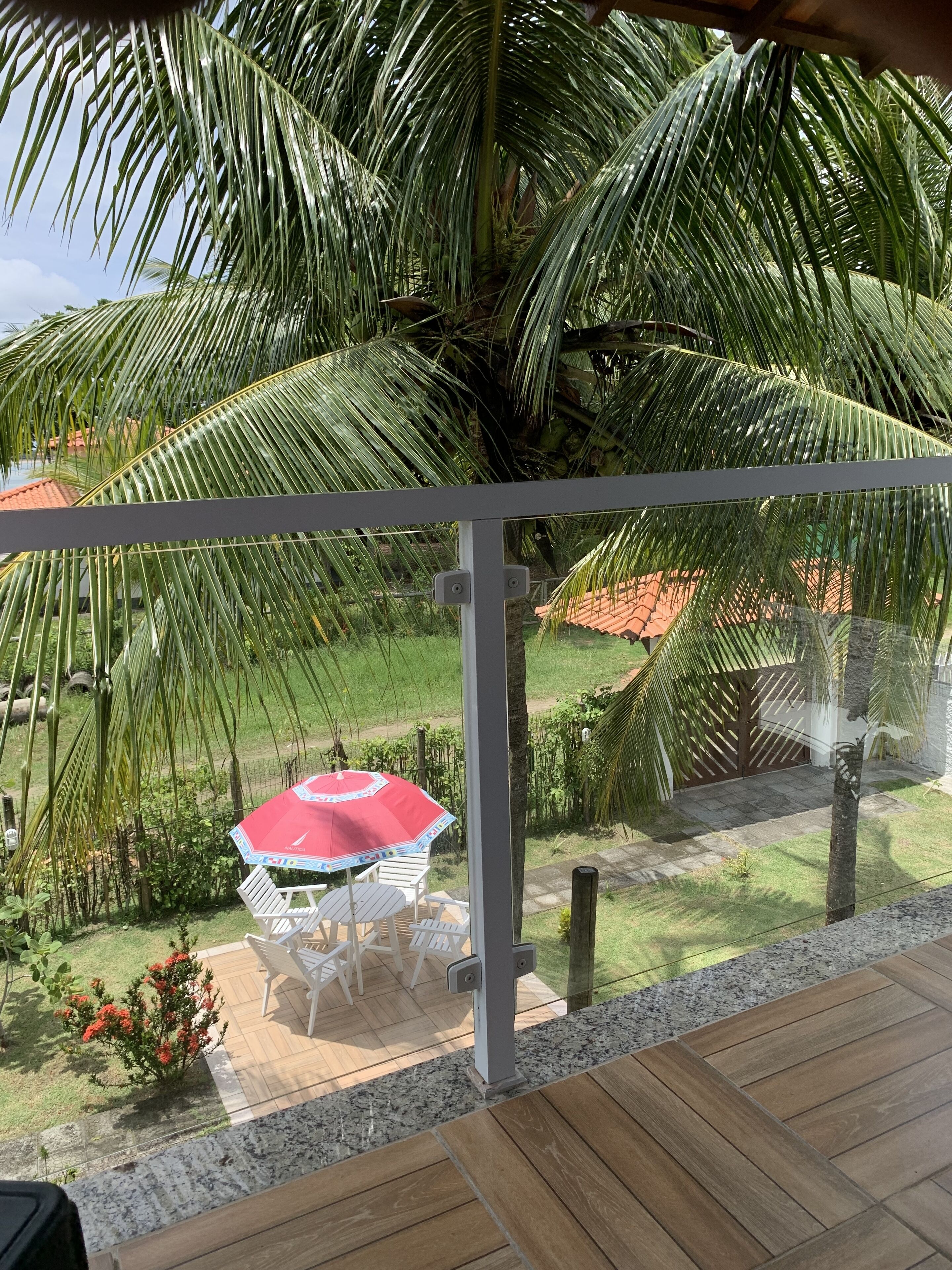 A balcony with a view of a garden, a palm tree, and a red umbrella table.