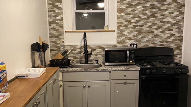 A kitchen with grey cabinets, a microwave, a stove, and a window with a hexagonal patterned backsplash.