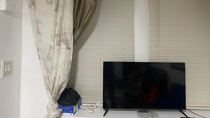 A room with a television on a stand, a window with blinds, a curtain with a Paris-themed print, and a power outlet on the wall.