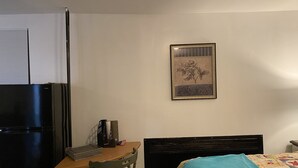 A bedroom with a bed, a desk with a chair, a refrigerator, a wall-mounted picture, and a window with blinds.