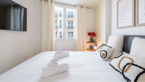 A hotel room with a bed, a TV mounted on the wall, a window with curtains, a bedside table with a vase of flowers, and framed artwork on the wall. - 2-Bedroom Apartment with Terrace and Side Sea View (Cannes)