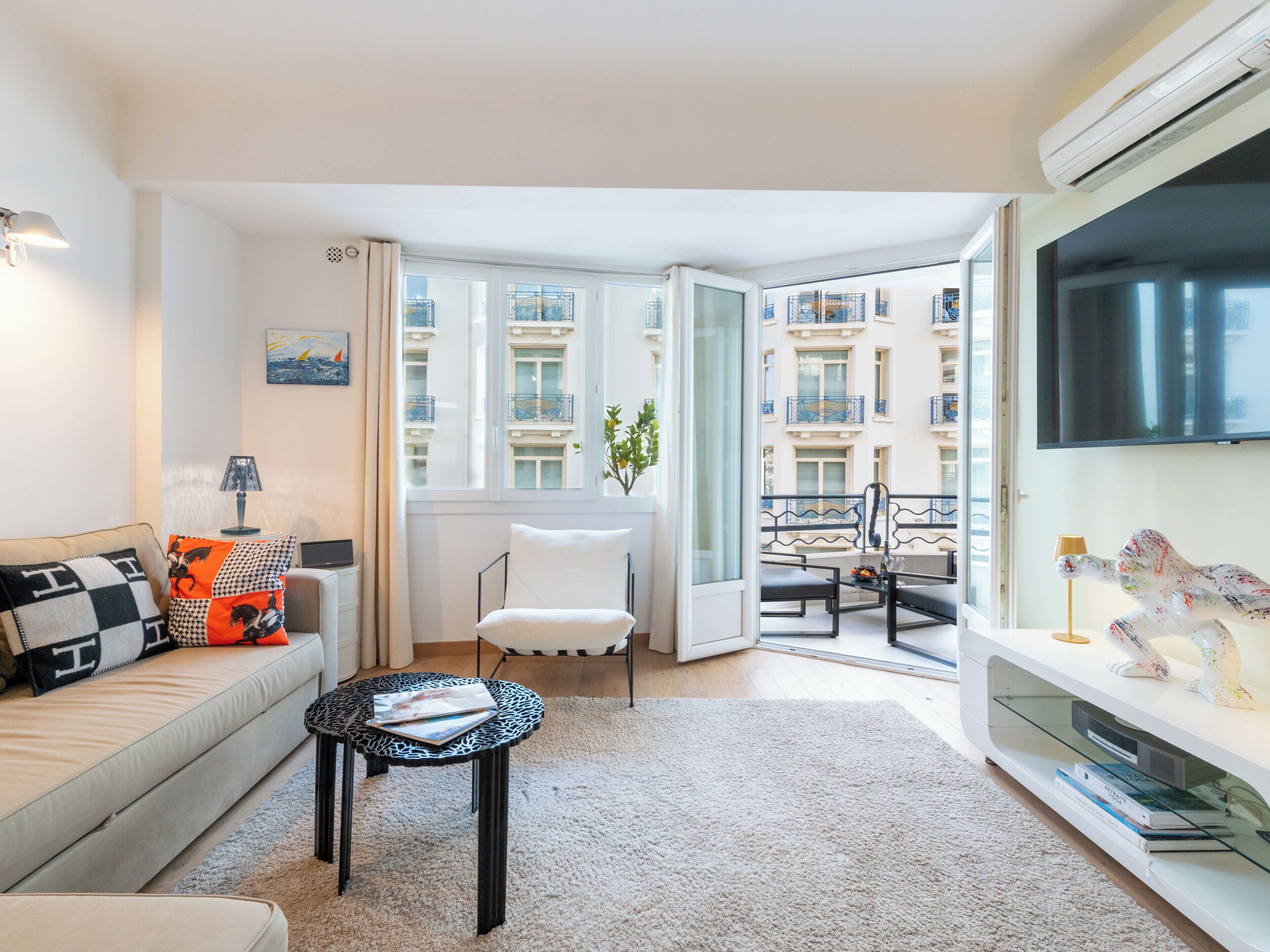 Ein modernes Wohnzimmer mit einer Couch, einem kleinen runden Tisch, einem Fernseher und einem Balkon mit Blick auf Gebäude.