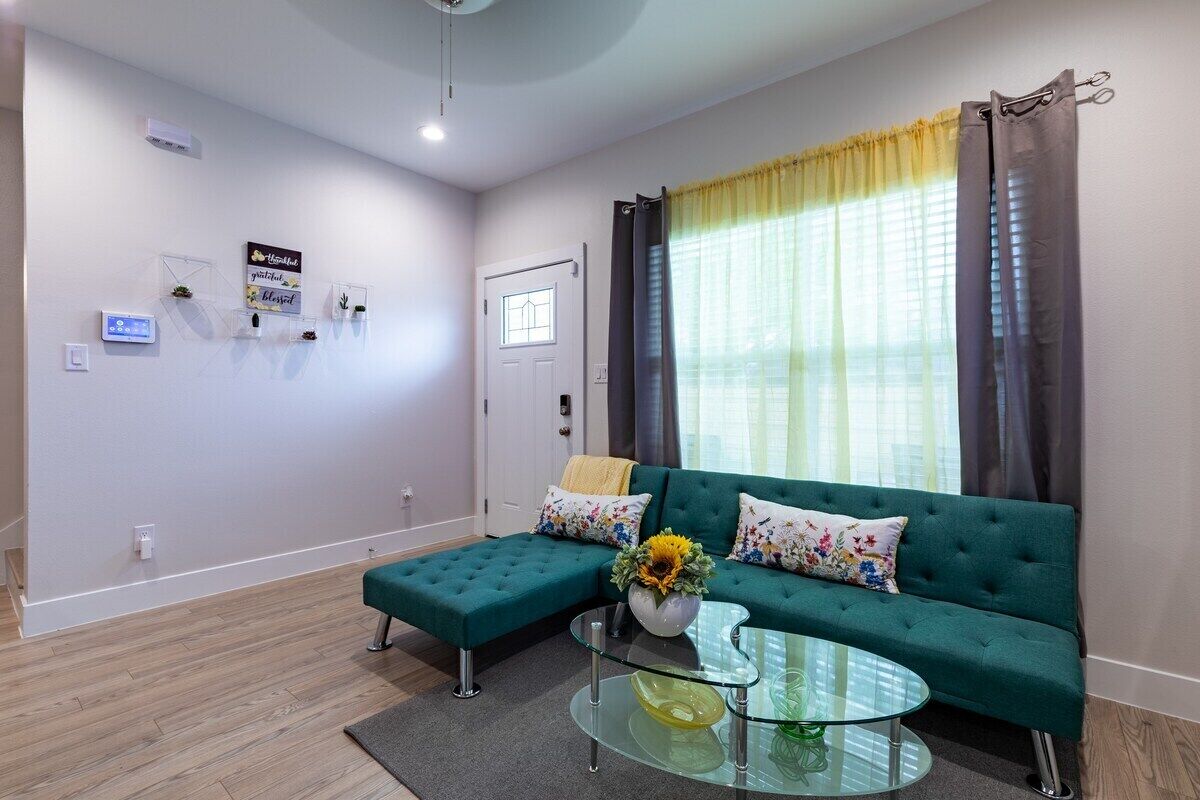 A modern living room with a green sofa, a glass coffee table, and a large window with curtains.