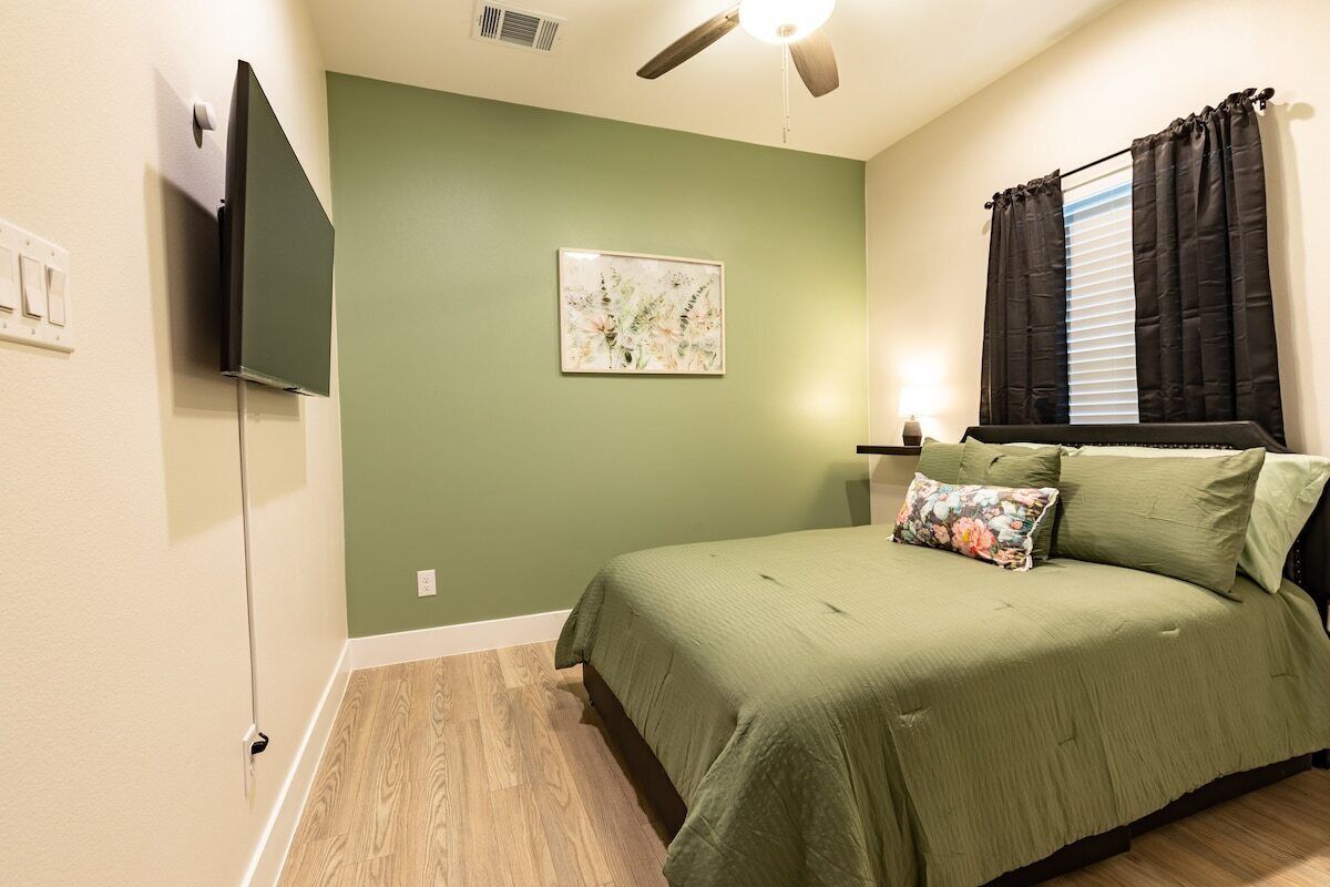 A bedroom with a bed, a ceiling fan, a wall-mounted TV, and a window with curtains.