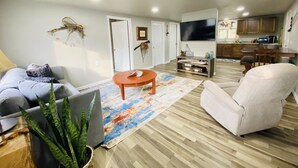 A modern living room with a grey sofa, a beige armchair, and a coffee table. There is a TV mounted on the wall and a kitchen area visible in the background. - Hooked On The River (Stites)
