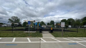 A playground with a wheelchair-accessible area, a fenced parking lot, and a cloudy sky.