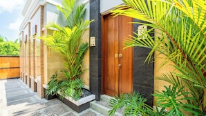 A modern building with a wooden door, surrounded by lush greenery and a stone pathway. - Villa Kamaratih (Legian)