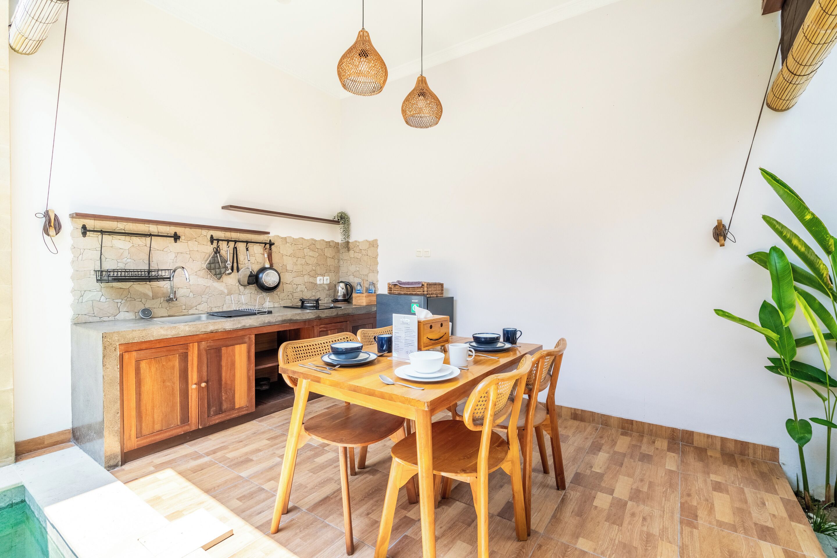 A dining area with a wooden table and chairs, a kitchen with stone backsplash and hanging utensils, and a hanging light fixture.