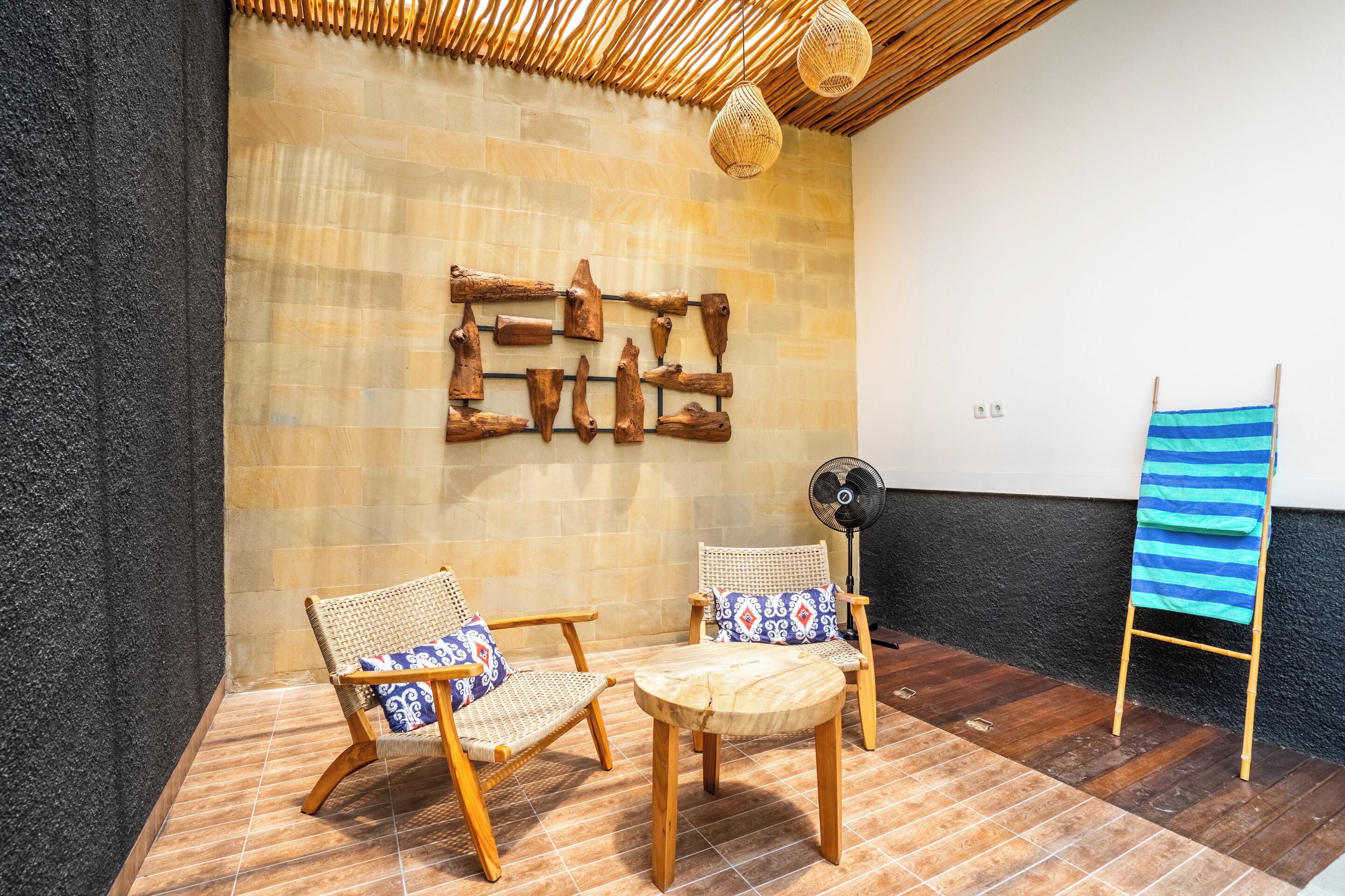 A room with two wooden chairs, a small round table, a wall-mounted fan, and a striped towel on a stand.