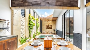 A dining area with a wooden table set for four, a small wooden side table, and a view of a swimming pool and outdoor seating area. - Villa Kamaratih (Legian)