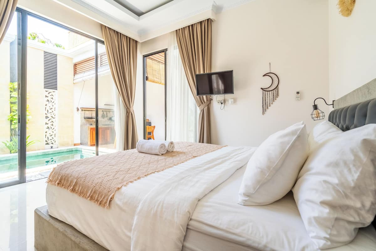 A hotel room with a large bed, a TV, a wall-mounted light, and a view of a pool.
