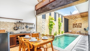 A modern kitchen with a stone backsplash, wooden dining table, and a pool area with a stone deck and lounge chairs. - Villa Kamaratih (Legian)