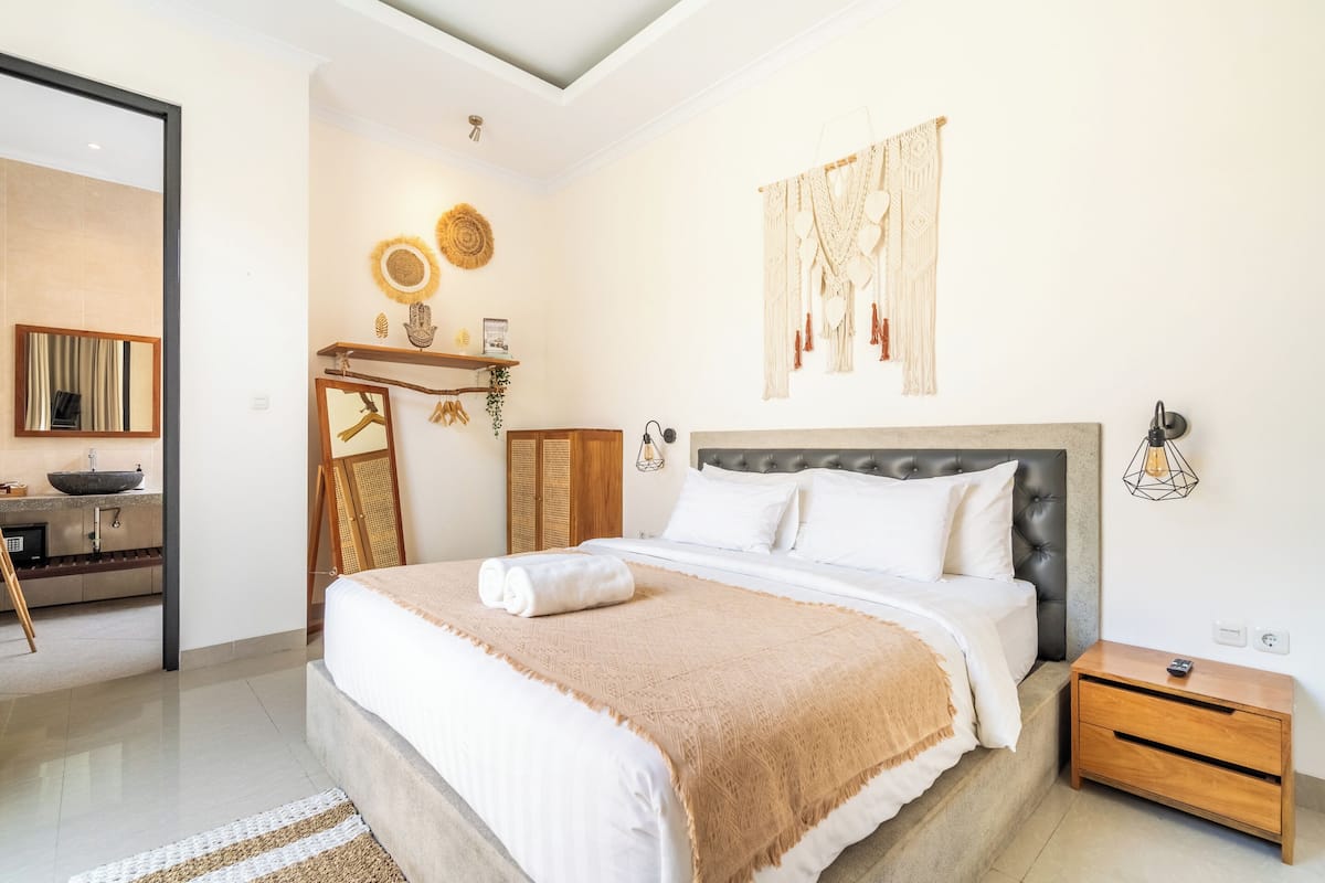 A modern bedroom with a large bed, a bedside table, a nightstand, a vanity with a mirror, and a wall-mounted macrame piece.