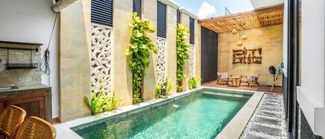 Una piscina con borde de piedra, rodeada por un muro de piedra con paneles decorativos y plantas trepadoras. Hay sillas de mimbre y una mesita cerca de la piscina.