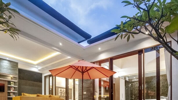 Un espace piscine extérieur moderne, doté d’un salon fermé par des baies vitrées, d’un canapé jaune et d’un parasol orange.