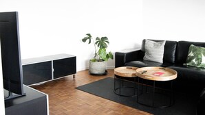Un salon moderne avec un canapé en cuir noir, deux petites tables en bois, une grande plante en pot et un téléviseur à écran plat.