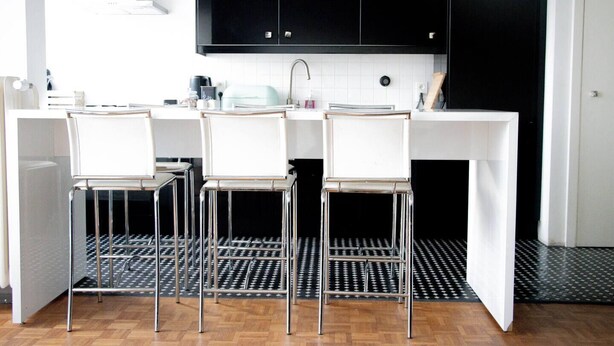 Une cuisine moderne avec trois tabourets de bar blancs, des meubles noirs et un plan de travail blanc.