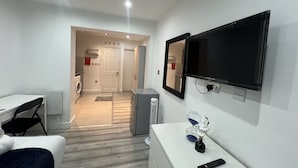 A hotel room with a bed, a desk, a chair, a TV, a mirror, and a small table with a vase and a remote control.