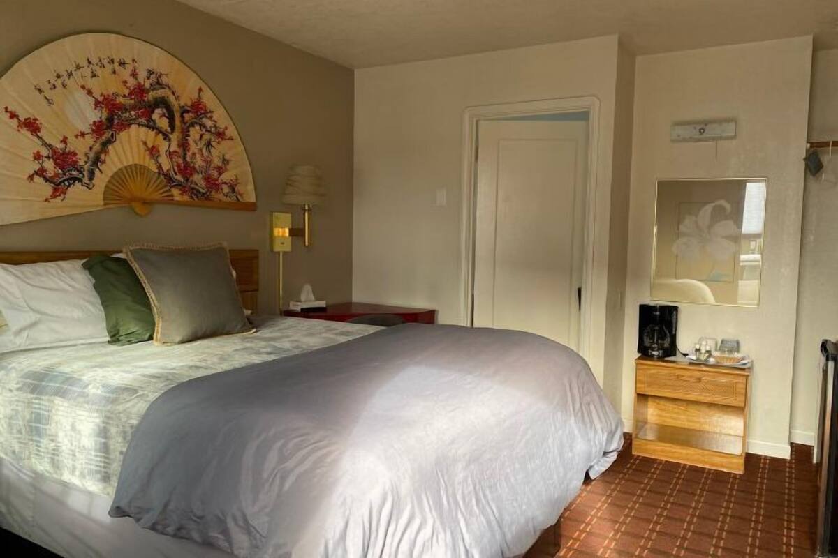 A bedroom with a large bed, a bedside table with a lamp, a wooden dresser, and a decorative fan on the wall.