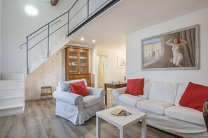 A modern living room with a staircase, white sofas with red cushions, a wooden cabinet, and a painting on the wall.