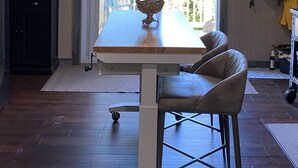 Un coin repas avec une table et des chaises en bois, un ventilateur de plafond, une horloge et une vue sur l’extérieur, visible à travers une porte vitrée.