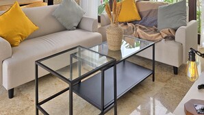 A modern living room with a glass coffee table, sofas with cushions, a kitchen area, and large windows with plants. - Villa Venta (Ungasan)