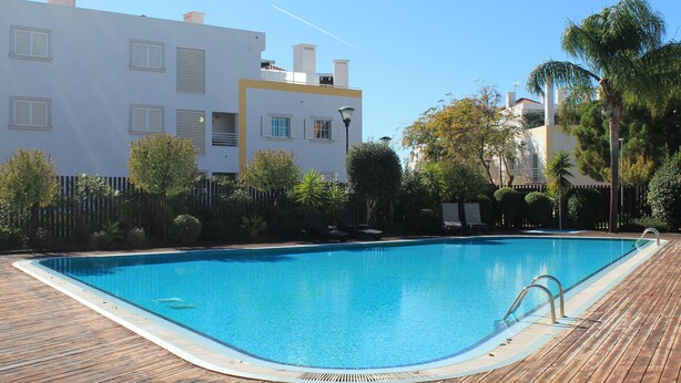 Une piscine rectangulaire entourée d’une terrasse en bois, le tout se fondant dans un décor composé d’immeubles modernes et de verdure.