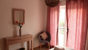 Une chambre à coucher avec un lit, une table de chevet, une chaise, un miroir et une fenêtre avec des rideaux.