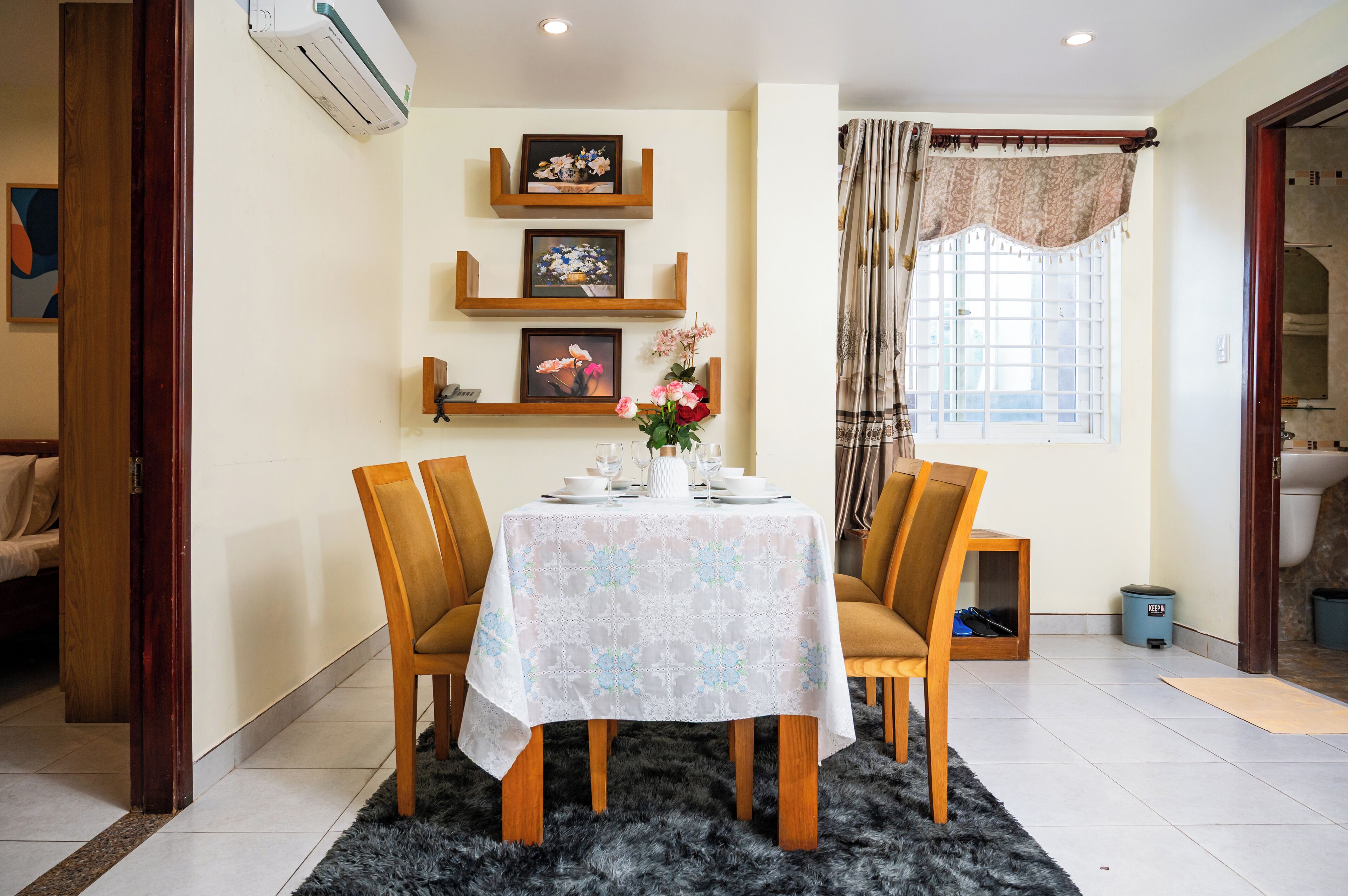 Un comedor con una mesa lista para cuatro comensales, flanqueada por sillas, un tapete decorativo y estanterías con cuadros.